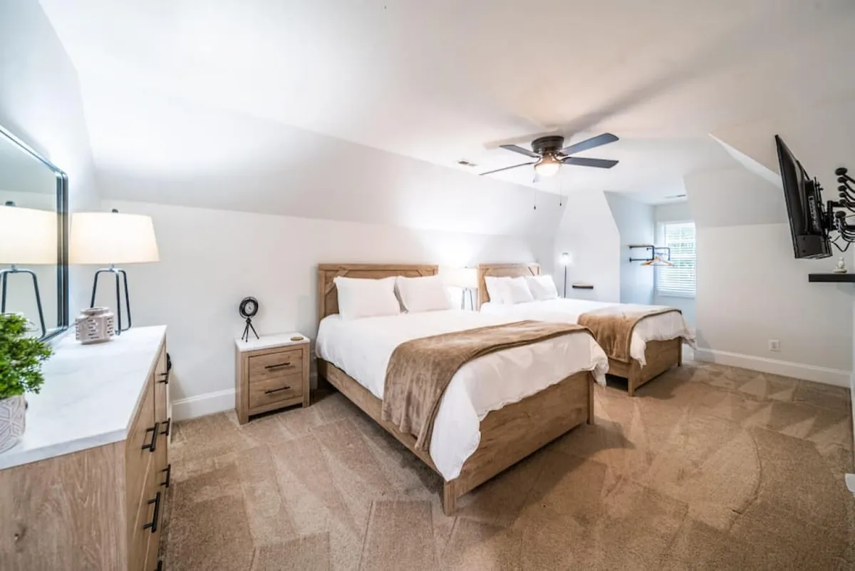 A serene bedroom with two queen beds dressed in soft linens, a modern dresser, a table lamp, and a flat-screen TV. The scene exudes comfort, encouraging restful nights and providing space for friends or colleagues traveling together.