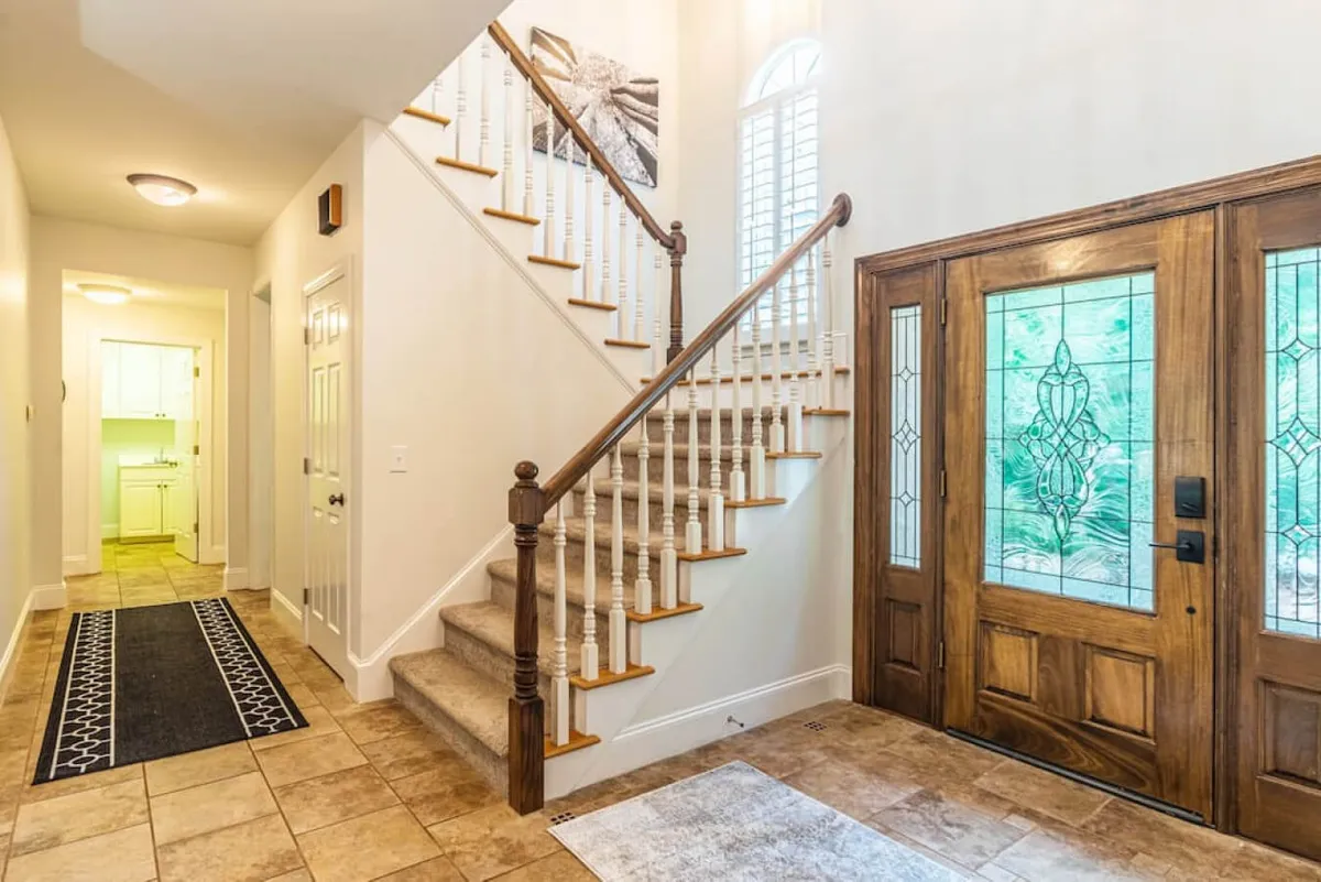 A bright, stylish entryway featuring a modern staircase, front door, decorative wall art, and an inviting area rug. The space feels open and welcoming, perfect for greeting friends or family as they arrive for their group getaway.
