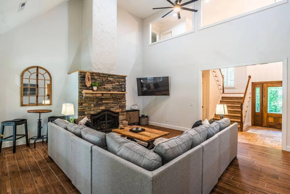 A spacious living room featuring a comfortable sectional sofa arranged around a wooden coffee table, with a stone fireplace as a central feature. Natural light pours in through large windows, and a TV is mounted on the wall, creating an inviting atmosphere for relaxation and group gatherings.