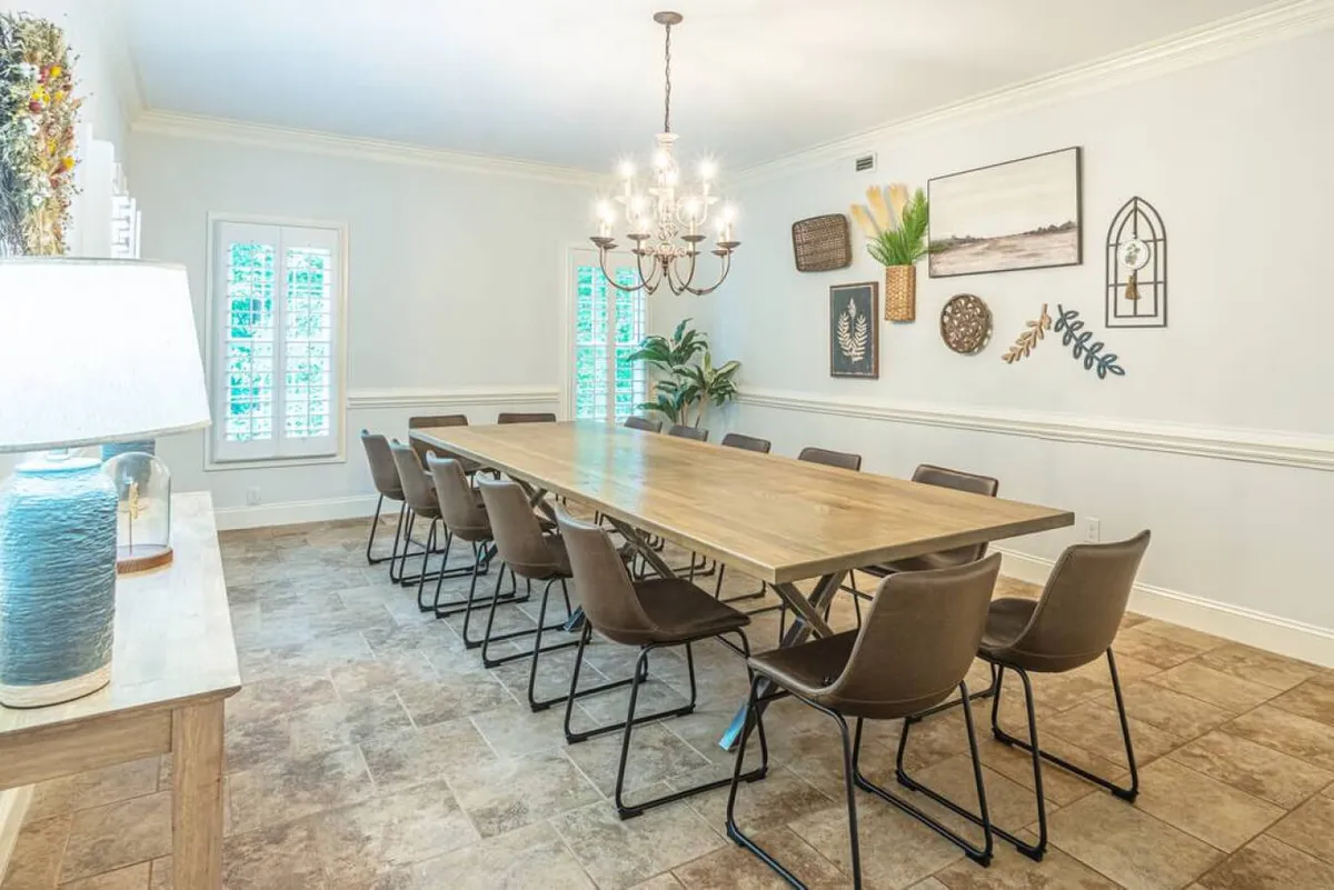 A spacious dining area centered around a long wooden table, illuminated by a chandelier, with decorative wall art and indoor plants adding warmth. The setting is ideal for family meals, group meetings, or celebrations, reflecting communal comfort and thoughtful design.