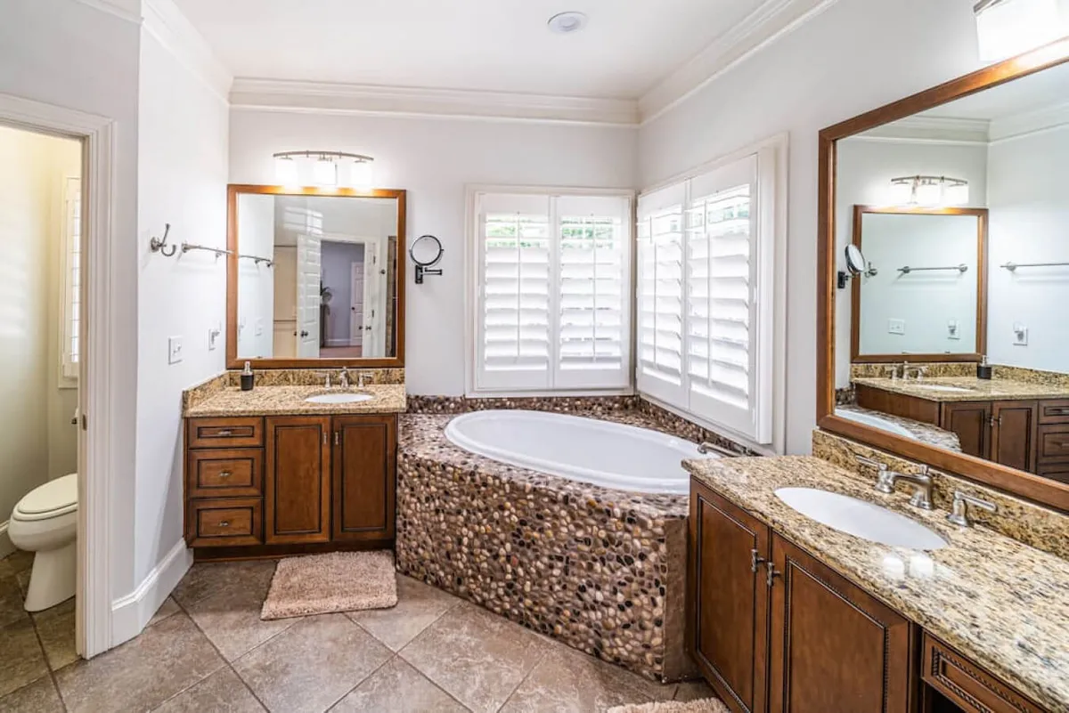 A spa-inspired bathroom with a luxurious soaking tub, granite countertops, large mirrors, and shuttered windows that bathe the space in natural light. The setting is serene, offering a peaceful place for relaxation and rejuvenation.