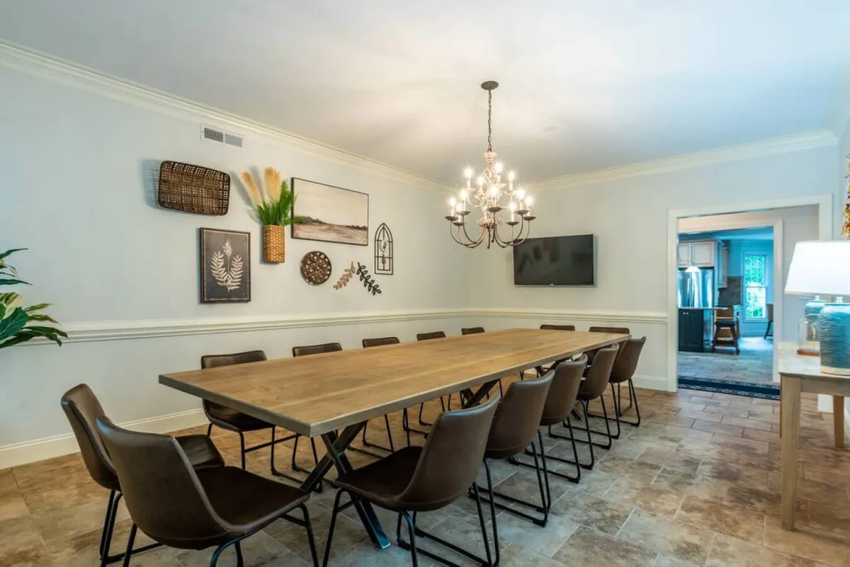 A long wooden dining table set beneath a stylish chandelier, surrounded by upholstered chairs. Decorative wall art and a flat-screen TV are visible, enhancing the multifunctional nature of the space. The room is bathed in warm light, projecting an atmosphere of togetherness and celebration.