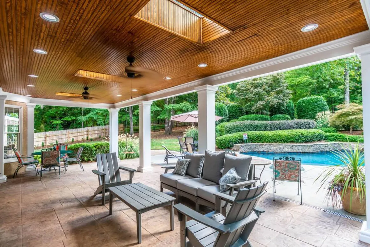 This image showcases an inviting outdoor seating area adjacent to a sparkling swimming pool, surrounded by a landscaped garden and covered by a ceiling with skylights. The space is designed for relaxation, entertaining guests, and enjoying meals or drinks outdoors, with views of both the pool and lush greenery.