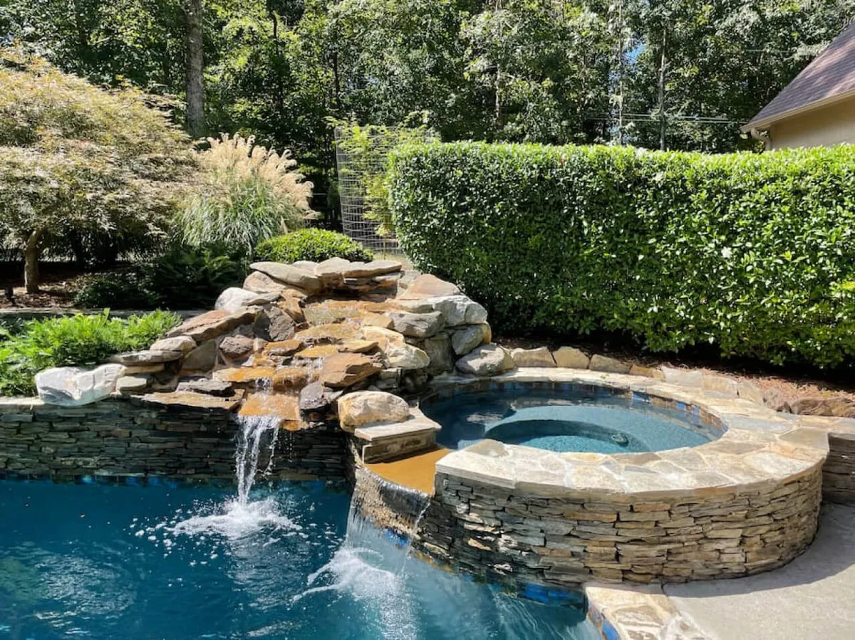 A tranquil exterior view showcasing a rock waterfall, jacuzzi, and lush greenery surrounding a gleaming blue pool. The scene evokes year-round relaxation and luxury, perfect for hot tub soaks or evening swims.