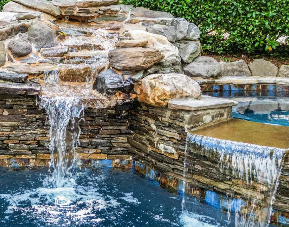 A serene exterior view of the villa’s swimming pool with a naturalistic waterfall and rock formations. The inviting pool area is surrounded by lush landscaping, offering both relaxation and entertainment spaces. The scene suggests ample room for group activities, social gatherings, or simply lounging poolside.