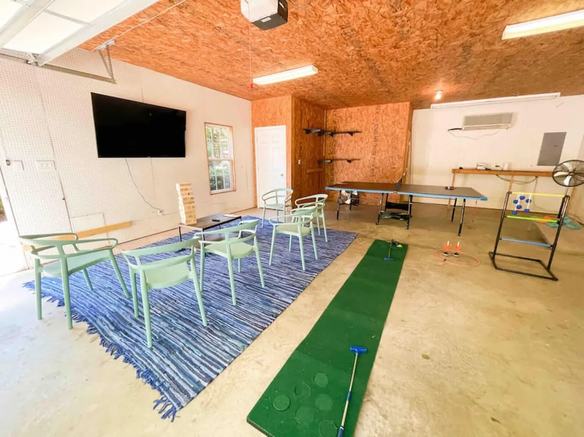 This interior shot features a playful space with green chairs, a ping pong table, a putting green for mini-golf, a rug with blue stripes, and a television. The room is designed for active recreation, fun competitions, and relaxed socializing, aligning with the garage game room description.
