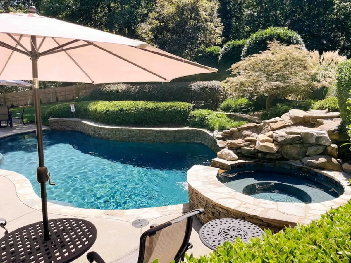 A serene exterior view of a backyard featuring a sparkling swimming pool, inviting hot tub, manicured gardens, and an outdoor seating area. The space is ideal for unwinding after a productive day or team-building events.