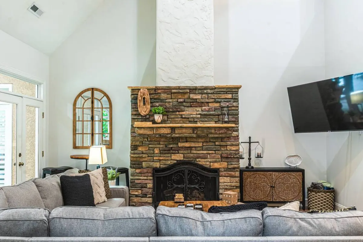 A cozy and stylish interior living space anchored by a stone fireplace, plush sofa, decorative mirror, and large television. The room invites guests to gather for movie nights, informal meetings, or game nights.