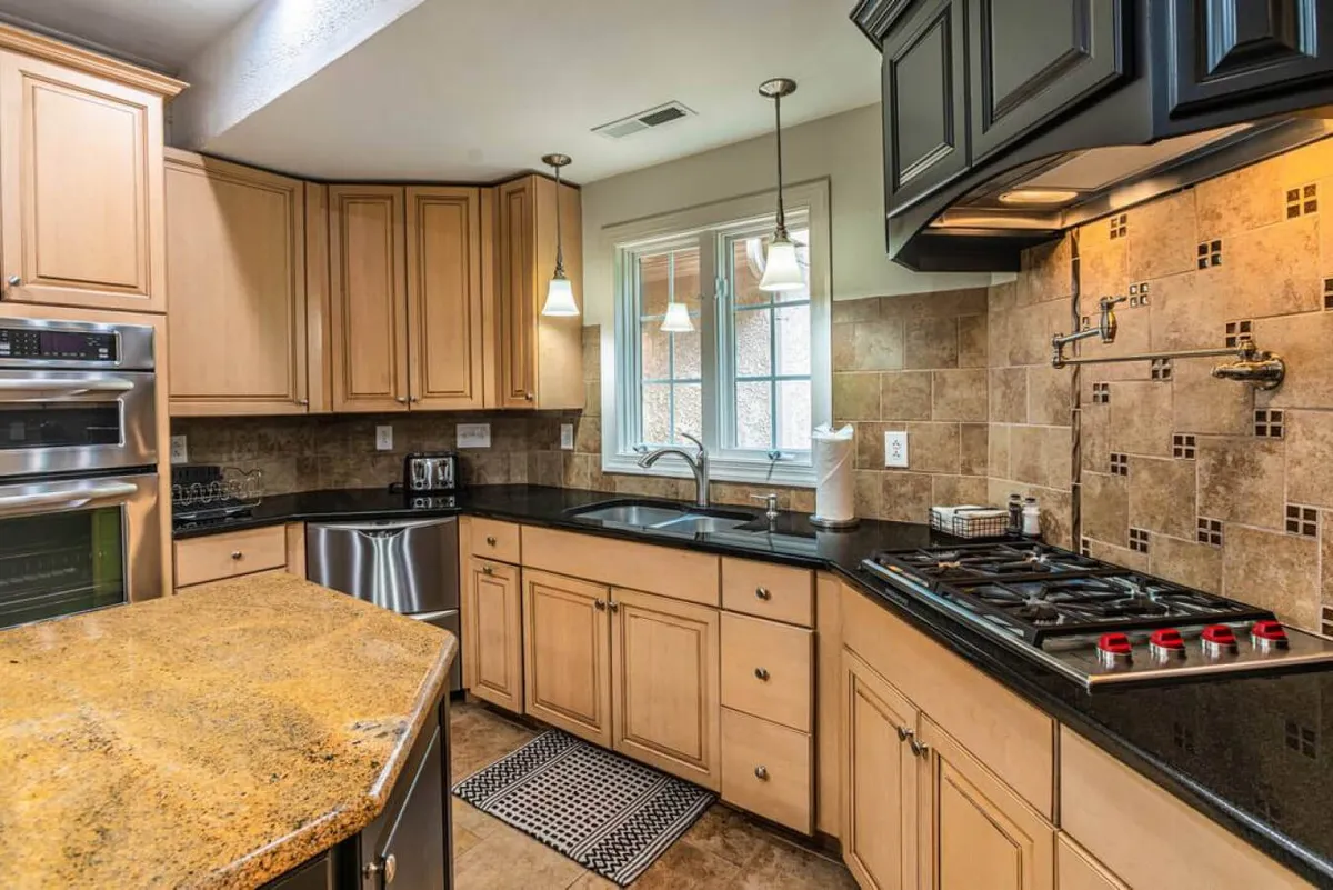 A modern kitchen featuring a gleaming granite countertop, stainless steel appliances, black cabinetry, and a sunlit window. The layout suggests ample cooking and prep space for group meals or catering to a crowd.